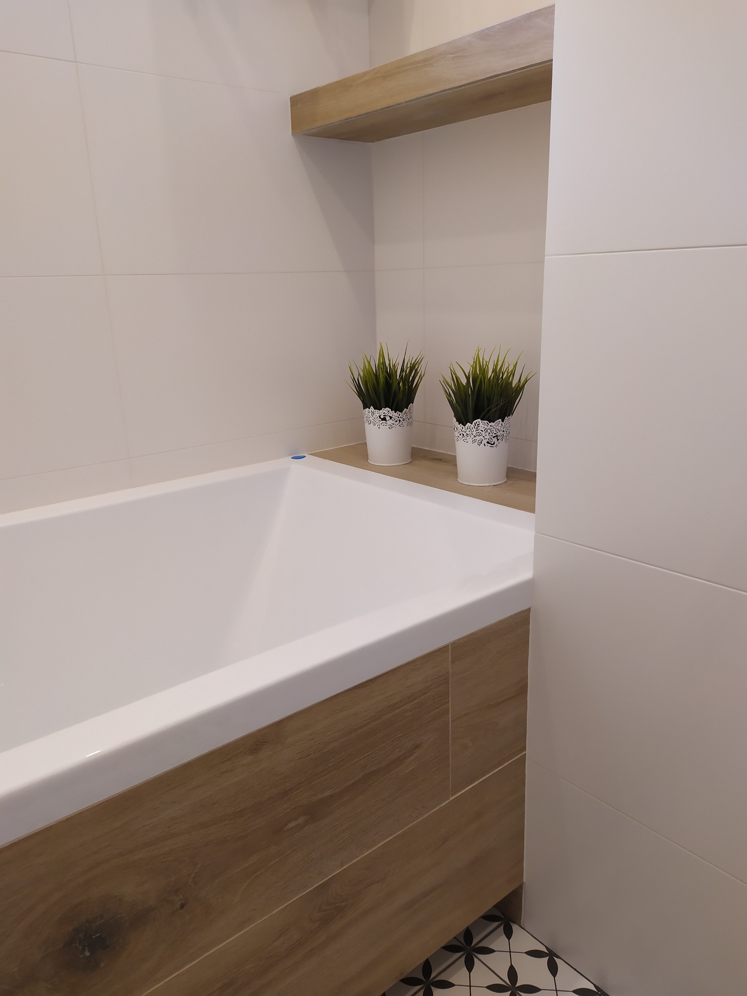 Nowoczesna łazienka z białą wanną obudowaną drewnopodobnymi płytkami, białe płytki ścienne i dekoracyjne rośliny w białych doniczkach na drewnianej półce.
