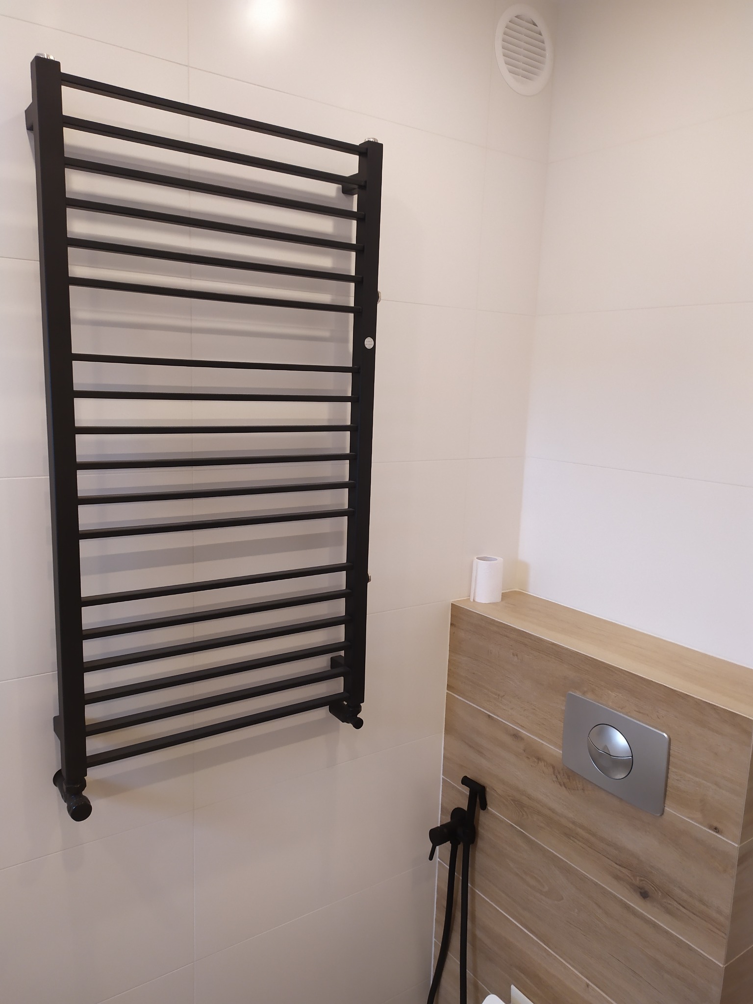 Czarny, drabinkowy grzejnik łazienkowy na białych, kwadratowych płytkach, z widocznym panelem WC w drewnopodobnej obudowie i czarnym bidetem.