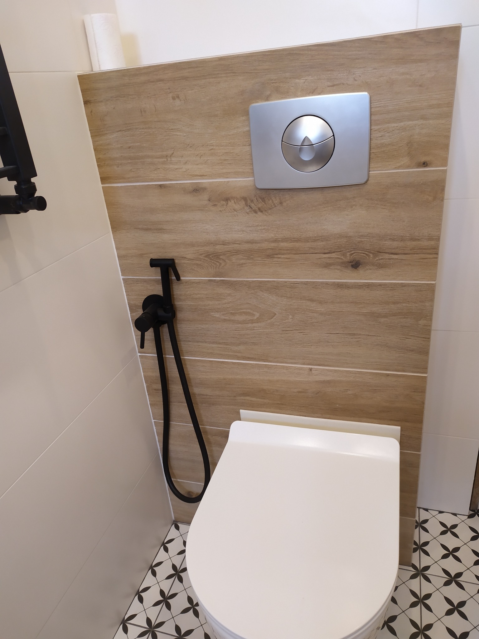 Podwieszana toaleta z drewnopodobną zabudową, chromowanym przyciskiem spłukującym i czarnym bidetem, na podłodze płytki w geometryczny wzór.