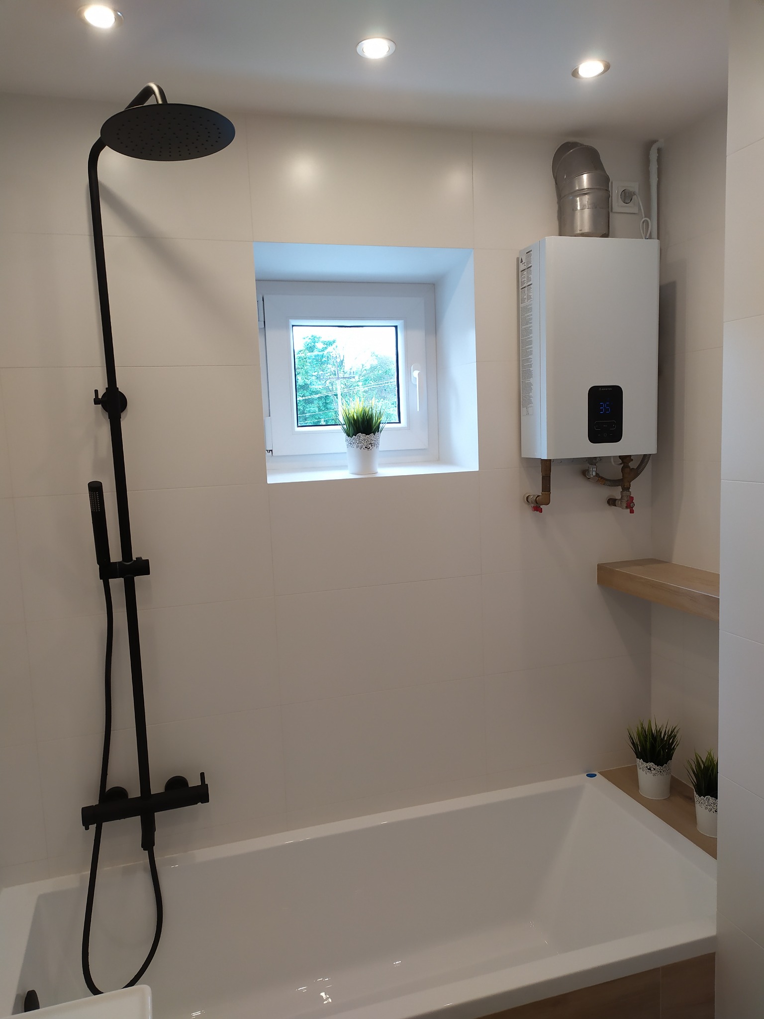 Biała łazienka z wanną, czarną armaturą prysznicową, podgrzewaczem wody i małym oknem z widokiem na zieleń.