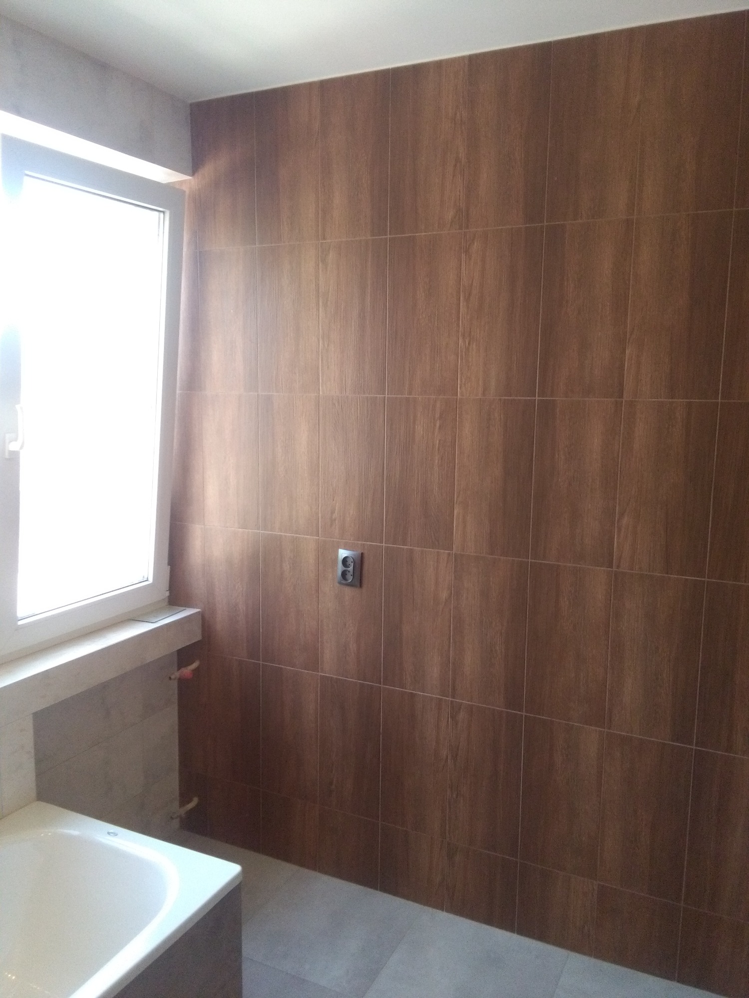 Ściana w łazience wyłożona brązowymi płytkami imitującymi drewno, z widocznym gniazdkiem elektrycznym, fragmentem okna z jasnym światłem oraz częścią wanny.