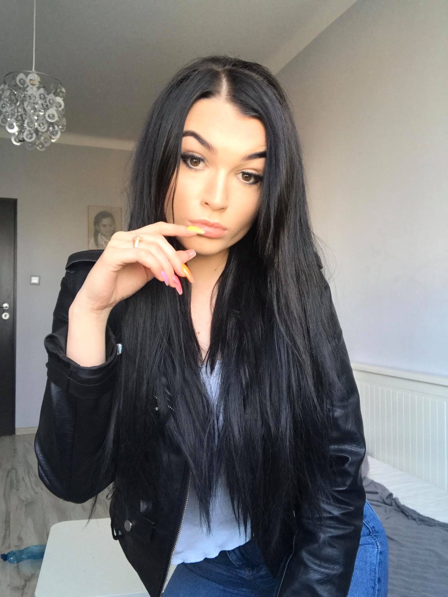 Portret młodej kobiety z długimi, czarnymi włosami, w czarnej skórzanej kurtce i niebieskich jeansach, pozującej w pokoju z białymi meblami i ozdobnym żyrandolem, z ręką uniesioną do ust i...