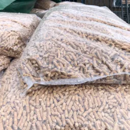 Stos worków z pelletem drzewnym, widoczne przez przezroczyste opakowanie, ułożonych na palecie pod zieloną plandeką.