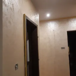 Beżowa ściana z dekoracyjną fakturą w korytarzu, widoczne zabezpieczone taśmą malarską ościeżnice drzwi, puszki elektryczne i fragment podłogi.