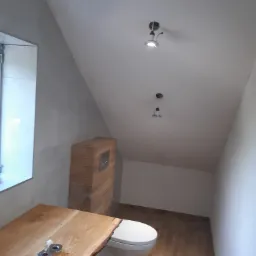 Wykończona łazienka na poddaszu ze skośnym sufitem i drewnianą podłogą, zabudowana toaleta z drewnianą obudową, drewniany blat umywalki z widocznymi krawędziami kory.