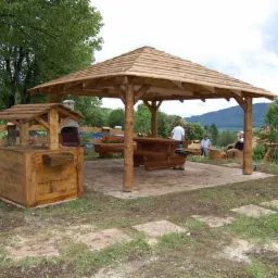 Drewniana pergola z kwadratowym dachem, studnia z daszkiem i drewniany stół z ławkami na zewnątrz, na tle zielonego wzgórza i gór.