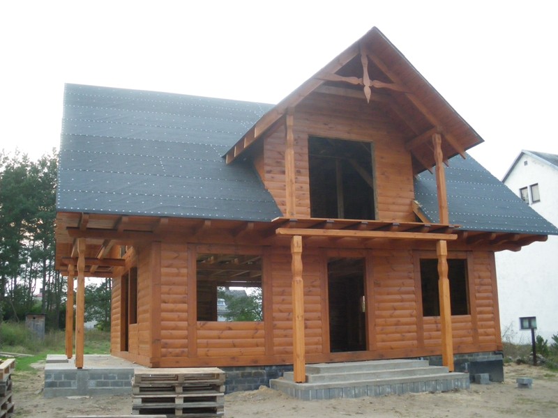 Surowy, dwukondygnacyjny dom z bali w trakcie budowy, z widoczną konstrukcją dachu pokrytą papą, podpartym balkonem i schodami wejściowymi z betonu.