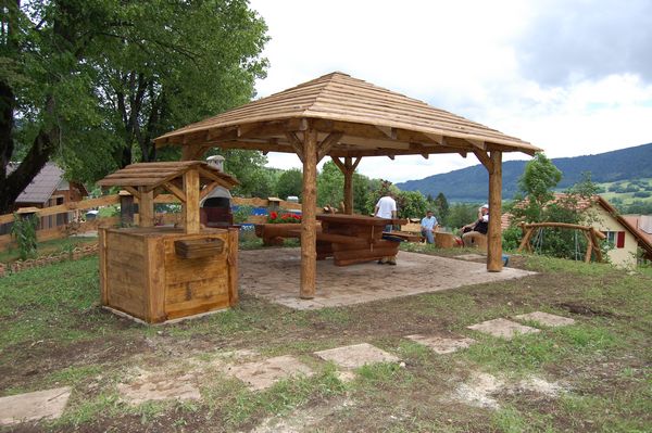 Drewniana pergola z kwadratowym dachem, studnia z daszkiem i drewniany stół z ławkami na zewnątrz, na tle zielonego wzgórza i gór.