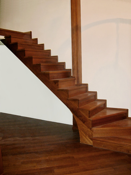 Schody z drewna egzotycznego z zakrętem, dopasowane do podłogi z parkietu w ciepłym odcieniu brązu, widoczne w jasnym wnętrzu.