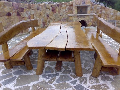 Rustykalny stół i ławki z litego drewna z widocznymi pęknięciami, ustawione na kamiennej posadzce z białym posypką, w tle murowany grill.