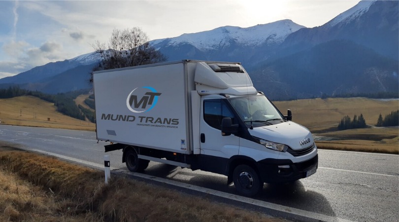 Biały samochód dostawczy marki Iveco z logo firmy transportowej na tle górskiego krajobrazu z ośnieżonymi szczytami i błękitnym niebem.