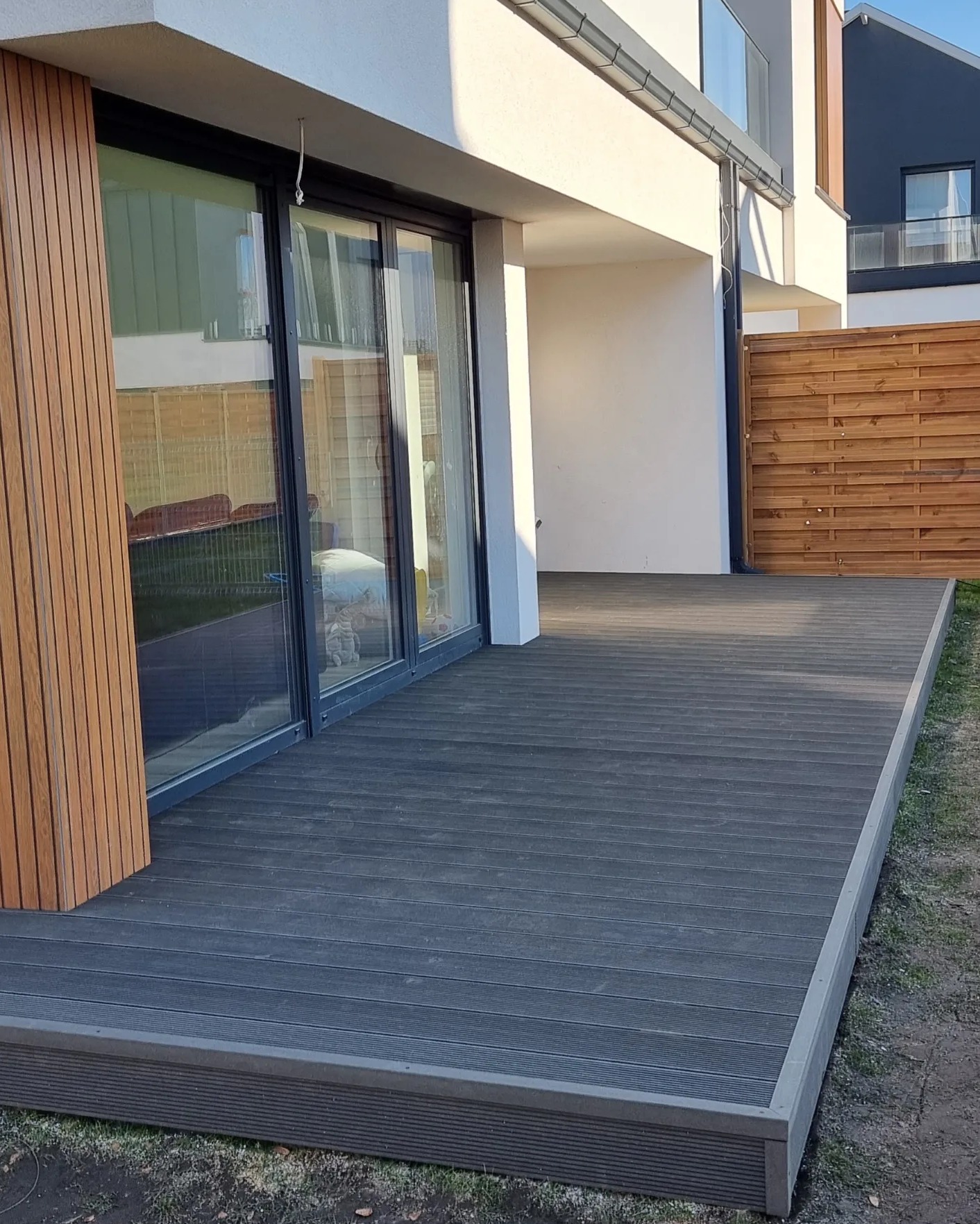 Nowoczesny taras z desek w kolorze grafitowym, przylegający do budynku z dużymi przesuwnymi drzwiami, widoczne elementy wykończeniowe i fragment drewnianego ogrodzenia w tle.