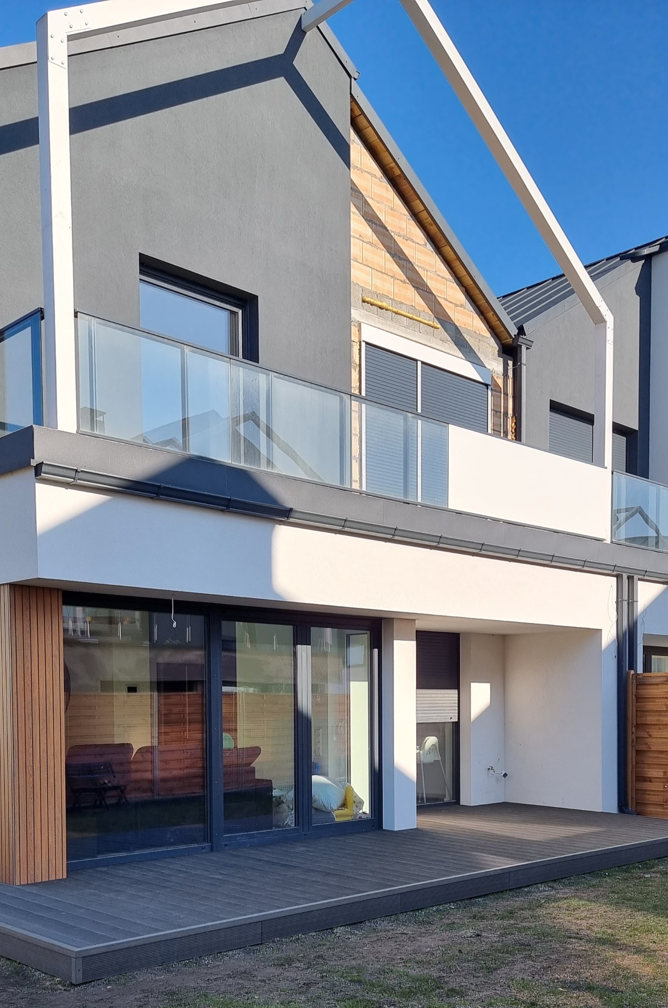 Nowoczesny dom z szarą elewacją, szklanymi balustradami na balkonie i tarasem z desek kompozytowych, widoczne elementy konstrukcji pergoli.