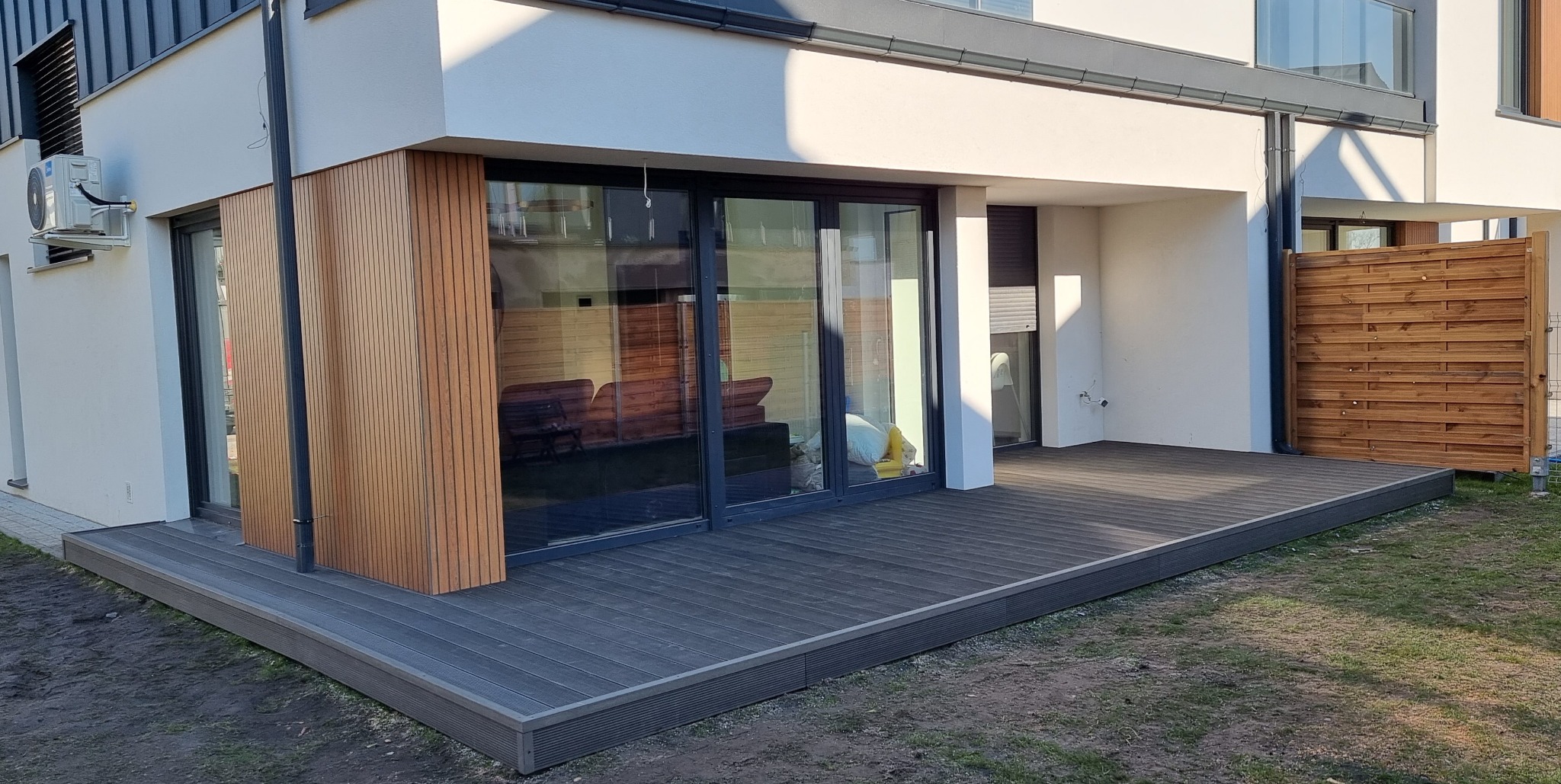 Nowoczesny taras z desek kompozytowych w kolorze grafitowym, przylegający do budynku z dużymi przesuwnymi drzwiami balkonowymi i elementami elewacji z drewnianych paneli. Widoczna drewniana ażurowa...