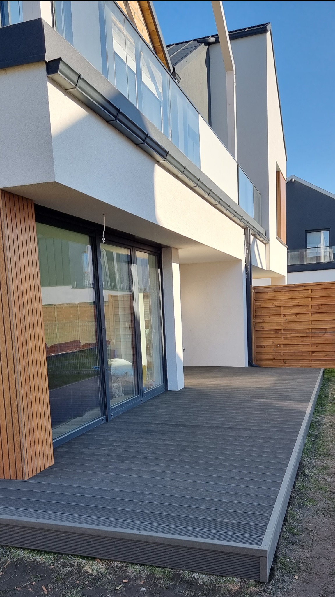 Nowoczesny taras z desek kompozytowych w kolorze grafitowym, przylegający do budynku z dużymi przesuwnymi oknami i drewnianą okładziną elewacyjną.