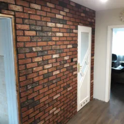 Ściana wykończona dekoracyjną cegłą w odcieniach czerwieni i brązu, sąsiadująca z białymi drzwiami z przeszklonymi elementami, widok na fragment korytarza z drewnianą podłogą i oświetleniem sufitowym.