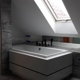 Nowoczesna wanna zabudowana w szarej łazience na poddaszu z oknem dachowym, wykończona płytkami imitującymi marmur.
