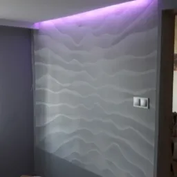 Szara ściana z dekoracyjną strukturą fal, podświetlona fioletowym światłem LED w górnej części, widoczne gniazdka elektryczne i fragment okna z firanką.