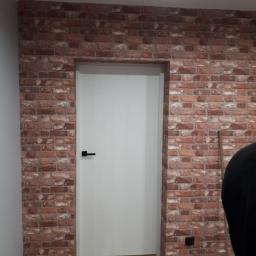 CeBRiS Sp. z o . o. - Ściana wykończona tapetą imitującą czerwoną cegłę z wkomponowanymi drzwiami z czarną klamką, widoczny fragment podłogi i czarnego ubrania oraz szczotka.