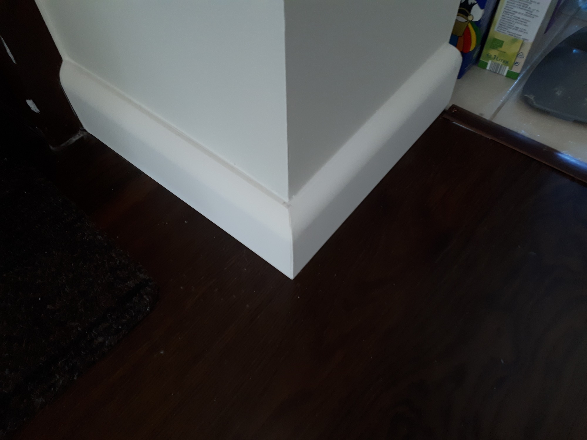 Szczegół narożnika ściany z białą listwą przypodłogową na tle ciemnej drewnianej podłogi i fragmentu dywanu.
