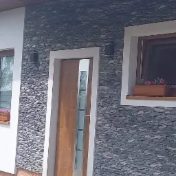 Elewacja domu z oknami w drewnianych ramach i nowoczesnymi drzwiami z przeszkleniami, wykończona kamieniem dekoracyjnym, z doniczkami z kwiatami pod oknami.