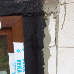 Uszczelnianie okna brązową ramą w Zambrów, widoczna czarna folia izolacyjna i świeży cement na białym murze.