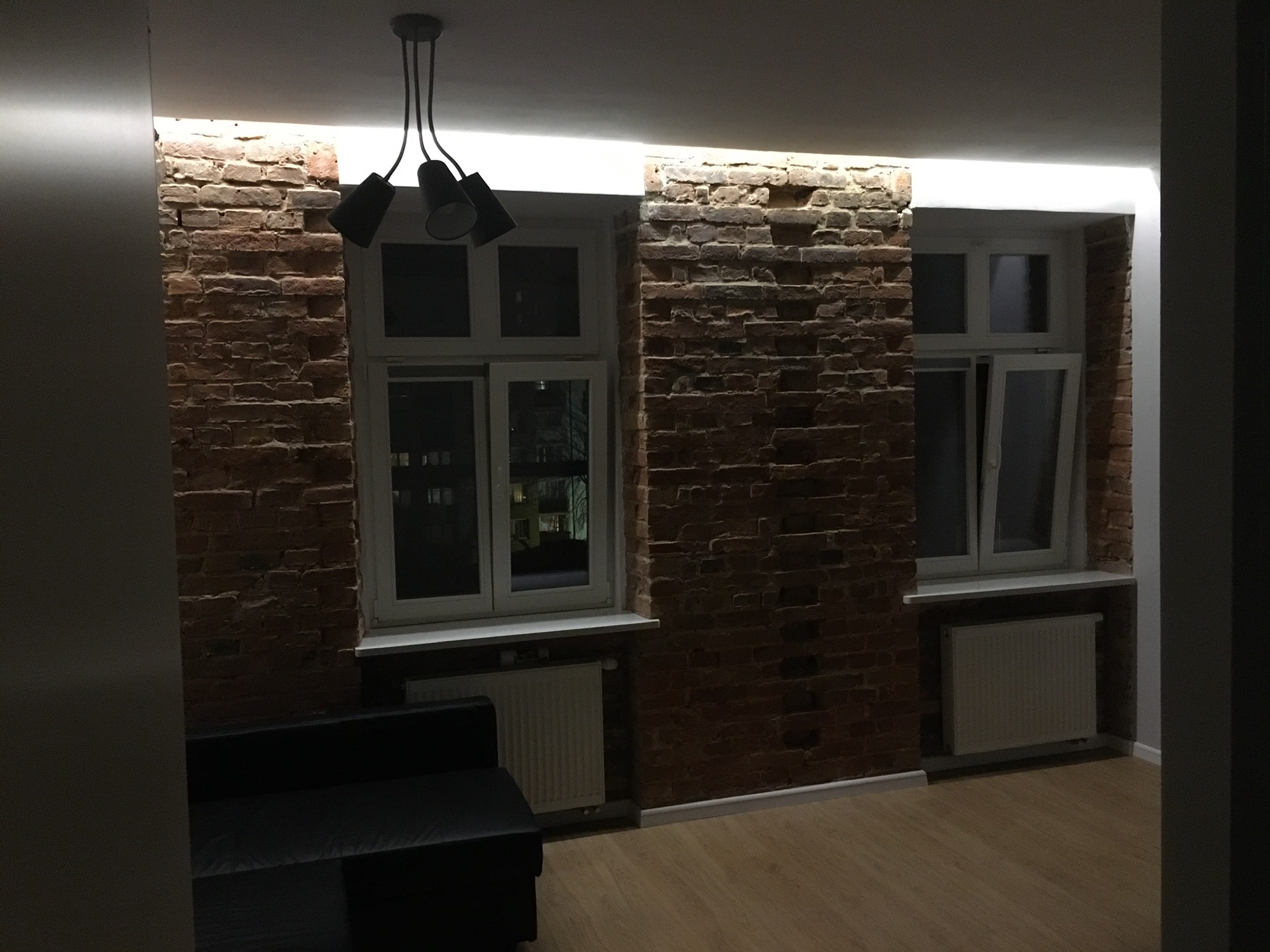 Ceglana ściana z dwoma oknami i grzejnikami podświetlona listwą LED, widoczny fragment sofy i lampy sufitowej.