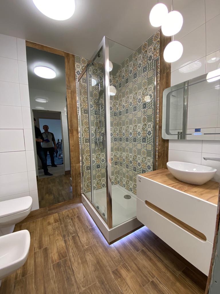 Nowoczesna łazienka z kabiną prysznicową, umywalką nablatową na szafce z drewnianym blatem, podświetleniem LED u dołu kabiny i drewnopodobną podłogą, widok z perspektywy wejścia.