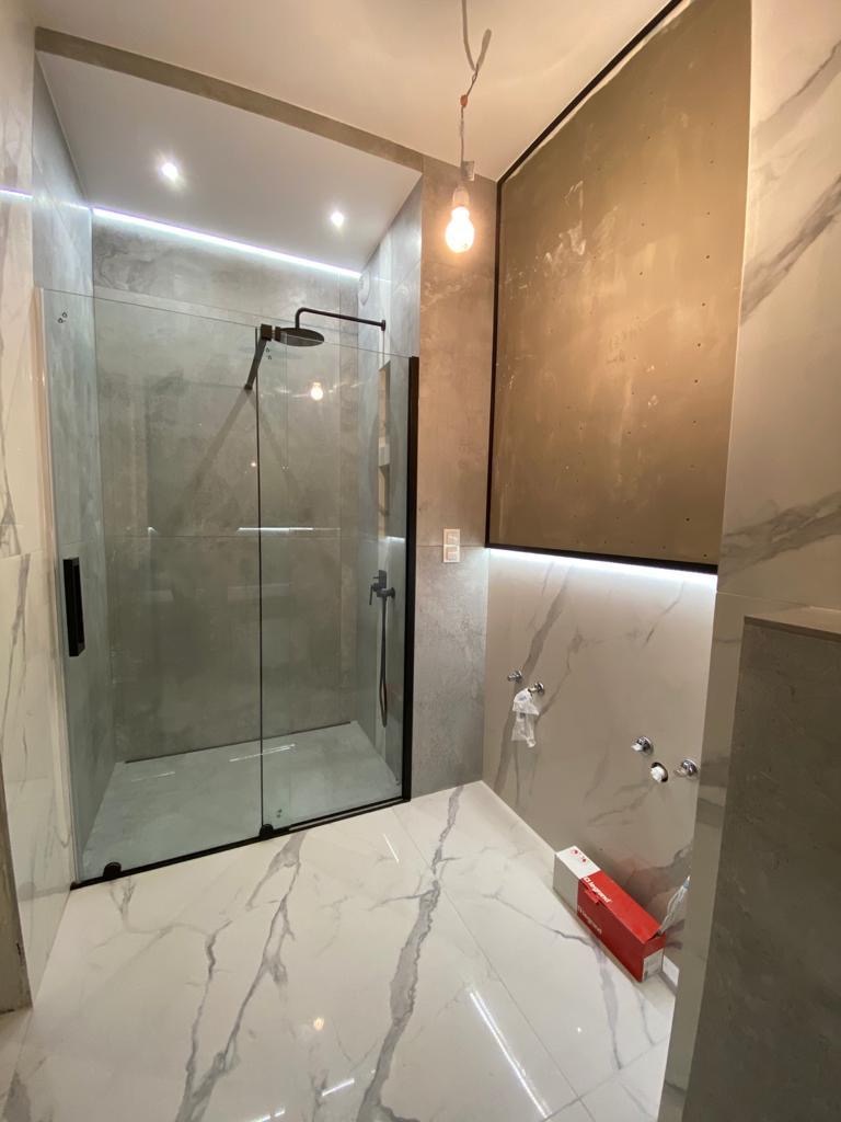 Wykończona łazienka z kabiną prysznicową typu walk-in, białe płytki imitujące marmur na podłodze i ścianach, surowa ściana przygotowana pod wykończenie, widoczna żarówka na kablu i pudełko...