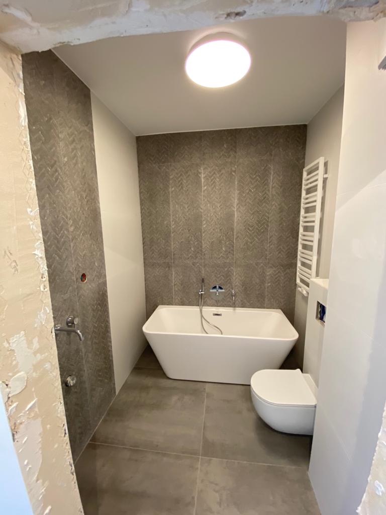 Nowoczesna łazienka z szarą mozaiką na ścianach, białą wanną wolnostojącą i podwieszaną toaletą, widoczny fragment ściany w stanie surowym przed wykończeniem.