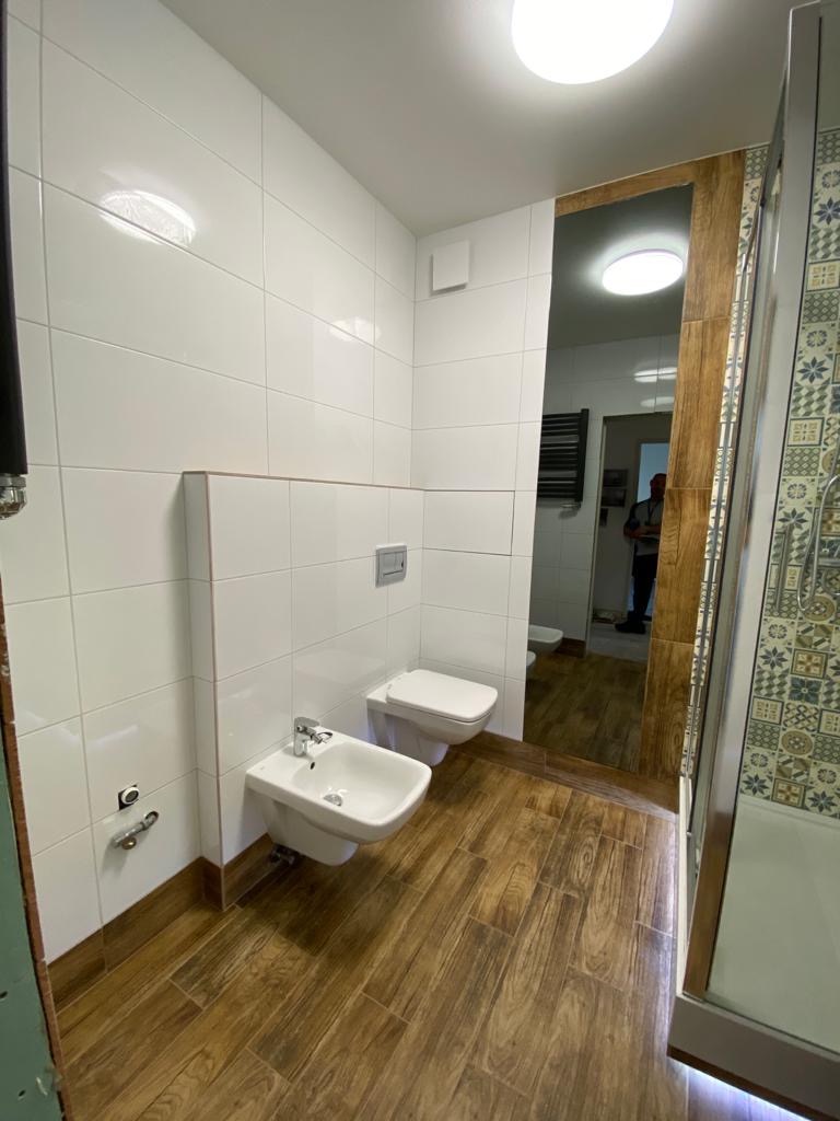 Wykończona łazienka z białymi, kwadratowymi płytkami na ścianach i drewnopodobną podłogą. Widoczna toaleta podwieszana, bidet i kabina prysznicowa z dekoracyjnymi płytkami. W tle widoczny fragment...