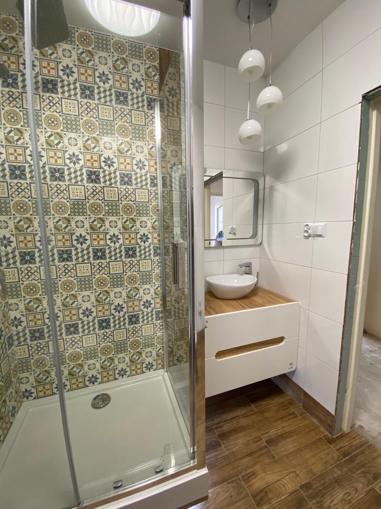 Nowoczesna łazienka z kabiną prysznicową z wzorzystymi płytkami, białą umywalką nablatową na szafce z drewnianym blatem, drewnopodobną podłogą i trzema wiszącymi lampami.