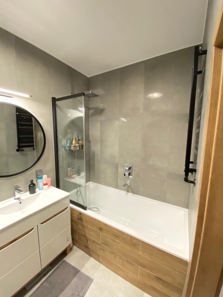 Nowoczesna łazienka z szarymi płytkami, białą wanną obudowaną drewnopodobnymi panelami, czarną kabiną prysznicową i okrągłym lustrem z podświetleniem LED nad białą szafką umywalkową.