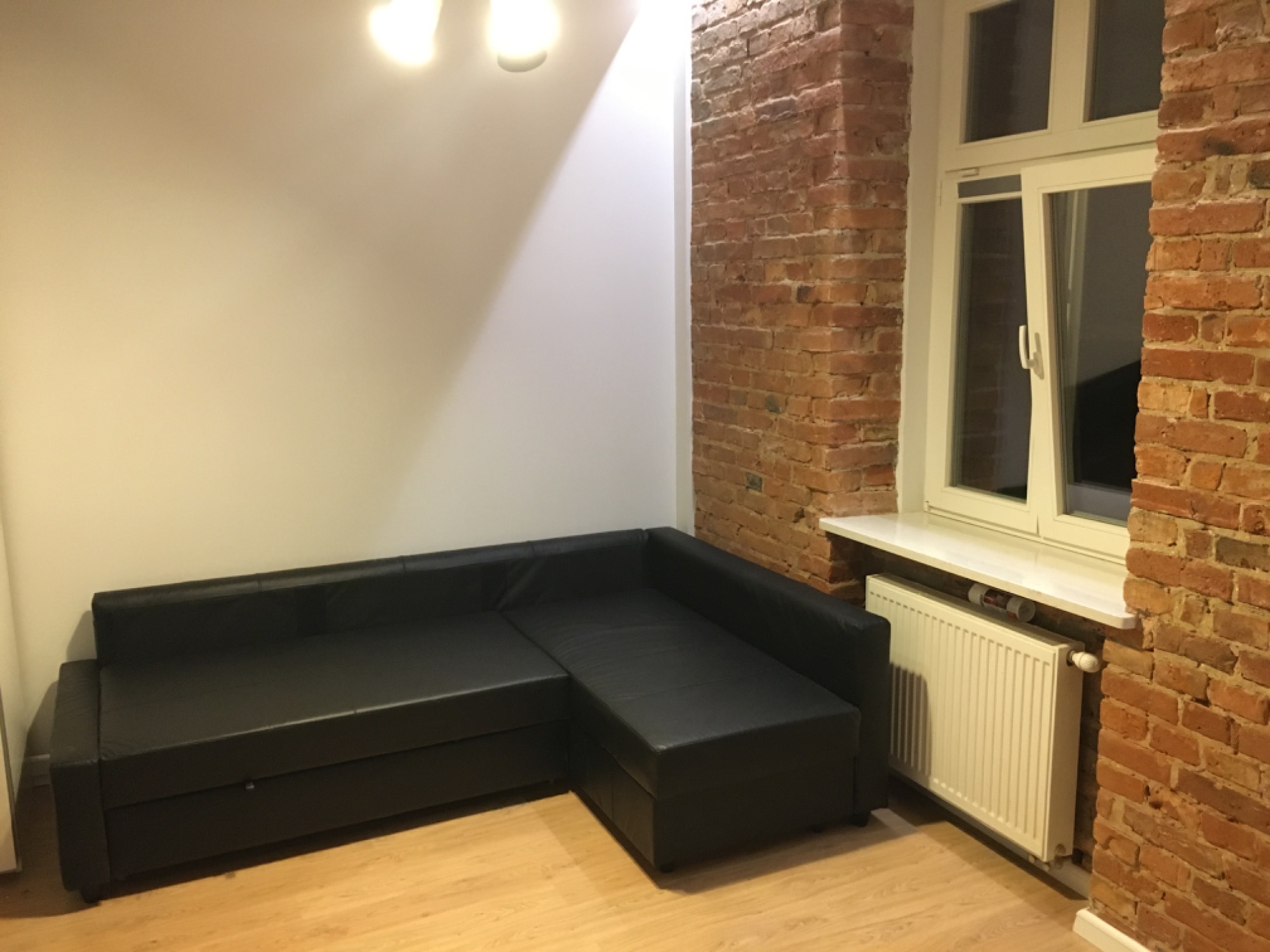 Ciemna, skórzana sofa narożna w rogu pokoju z białą ścianą i ścianą z czerwonej cegły z oknem; widoczny biały grzejnik pod oknem.