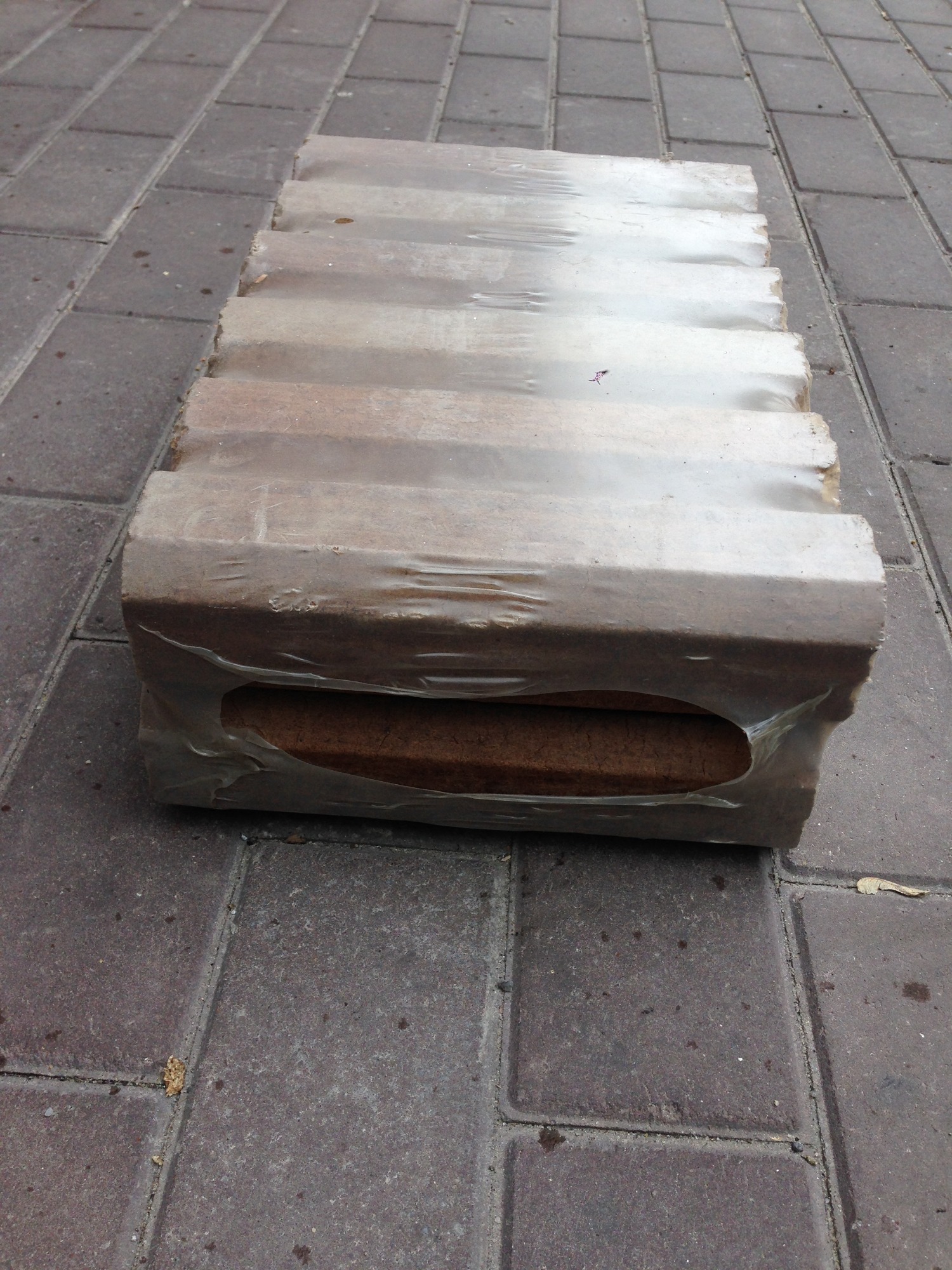 Paczka brykietu drzewnego owinięta w folię, leżąca na szarej kostce brukowej.