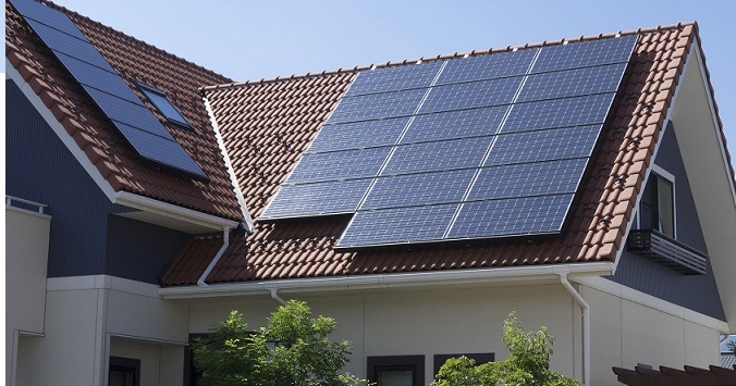 Panele słoneczne na dachu domu jednorodzinnego z czerwoną dachówką, widoczne okno dachowe i fragment elewacji.