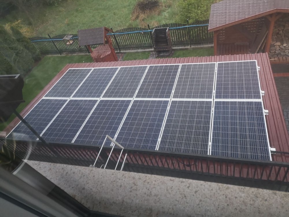 Widok z góry na panele słoneczne zamontowane na dachu budynku z czerwonej blachy trapezowej, w tle widoczny ogród z altaną i grillem.