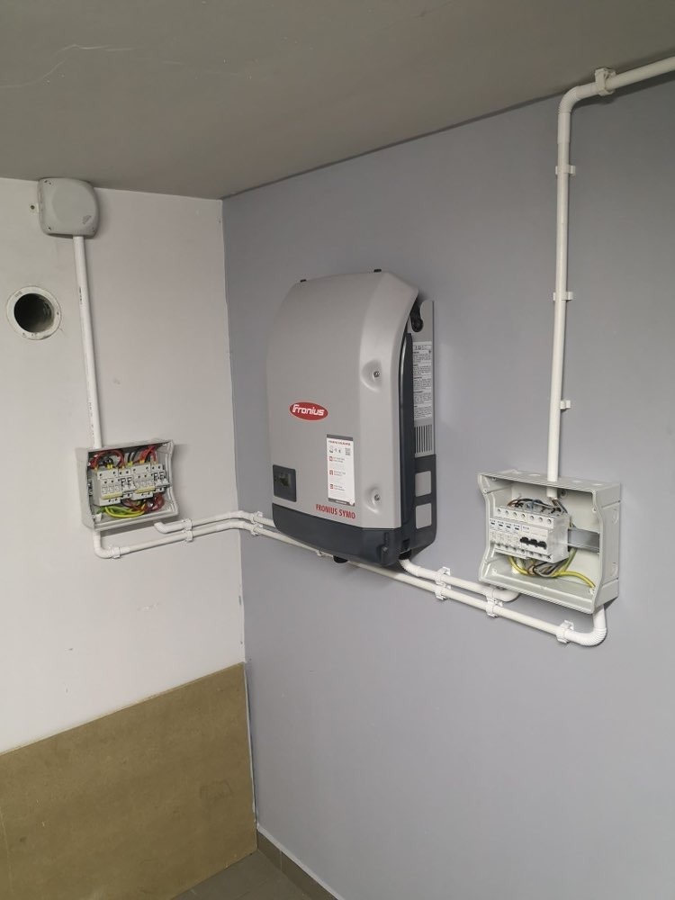 Szary inwerter Fronius Symo zamontowany na szarej ścianie w pomieszczeniu, połączony białymi rurkami z dwiema szarymi skrzynkami elektrycznymi.