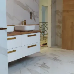 Nowoczesna łazienka w trakcie wykańczania, białe marmurowe płytki na ścianach i podłodze, biała szafka pod umywalką z drewnianym blatem i złotymi uchwytami, widoczne niedokończone prace instalacyjne.