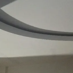 Kremowy sufit z zaokrąglonym obniżeniem, widoczne mocowanie lampy wiszącej, ściana w tle w odcieniach beżu.