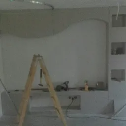 Ściana w trakcie wykańczania z dekoracyjnym łukiem i wnękami, widoczna drabina, narzędzia i laserowy poziom, surowy stan pomieszczenia.