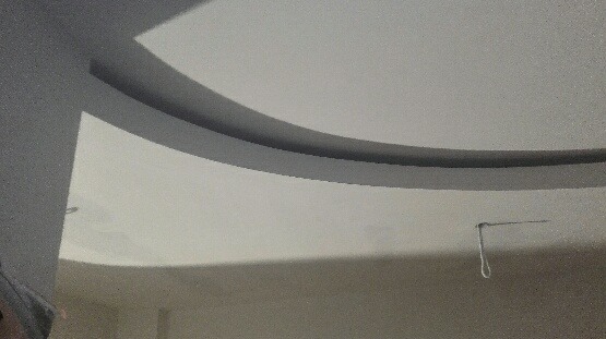 Kremowy sufit z zaokrąglonym obniżeniem, widoczne mocowanie lampy wiszącej, ściana w tle w odcieniach beżu.