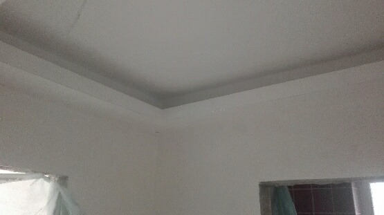 Pomieszczenie po malowaniu na biało, widoczny fragment sufitu z listwą dekoracyjną przy ścianie, okno zabezpieczone folią malarską.