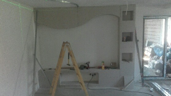 Ściana w trakcie wykańczania z dekoracyjnym łukiem i wnękami, widoczna drabina, narzędzia i laserowy poziom, surowy stan pomieszczenia.