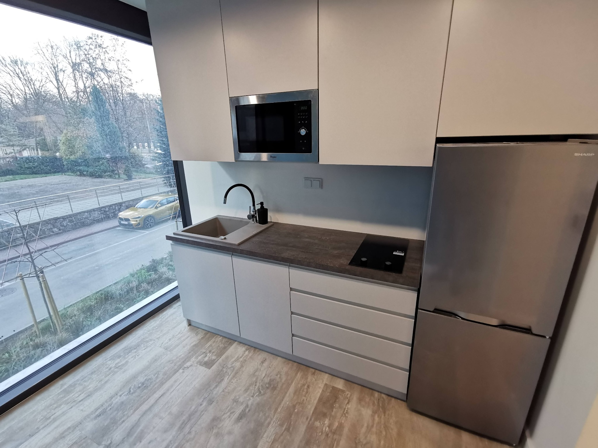 Nowoczesna zabudowa kuchenna w odcieniach szarości z widokiem na ulicę przez panoramiczne okno, obejmująca szafki wiszące, zintegrowaną mikrofalówkę, zlew, płytę indukcyjną i lodówkę Sharp.