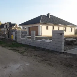 Niski murek z szarego betonu architektonicznego, stanowiący część ogrodzenia posesji z nowoczesnym domem jednorodzinnym w tle.