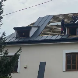 Ekipa dekarzy montuje blachodachówkę na dachu domu z lukarnami, widoczny fragment konstrukcji więźby dachowej.