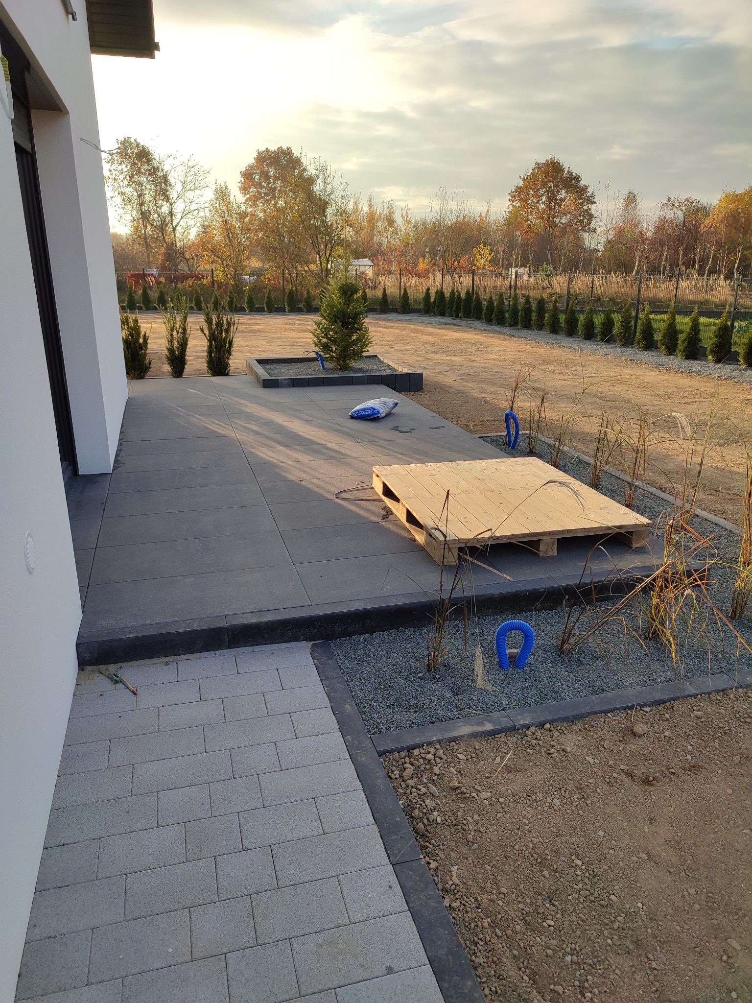 Nowoczesny taras z szarych płyt betonowych, obrzeża z ciemnego kamienia, rabaty z ozdobnym żwirem i trawami, w tle rząd tuj i jesienne drzewa, paleta drewniana na tarasie.