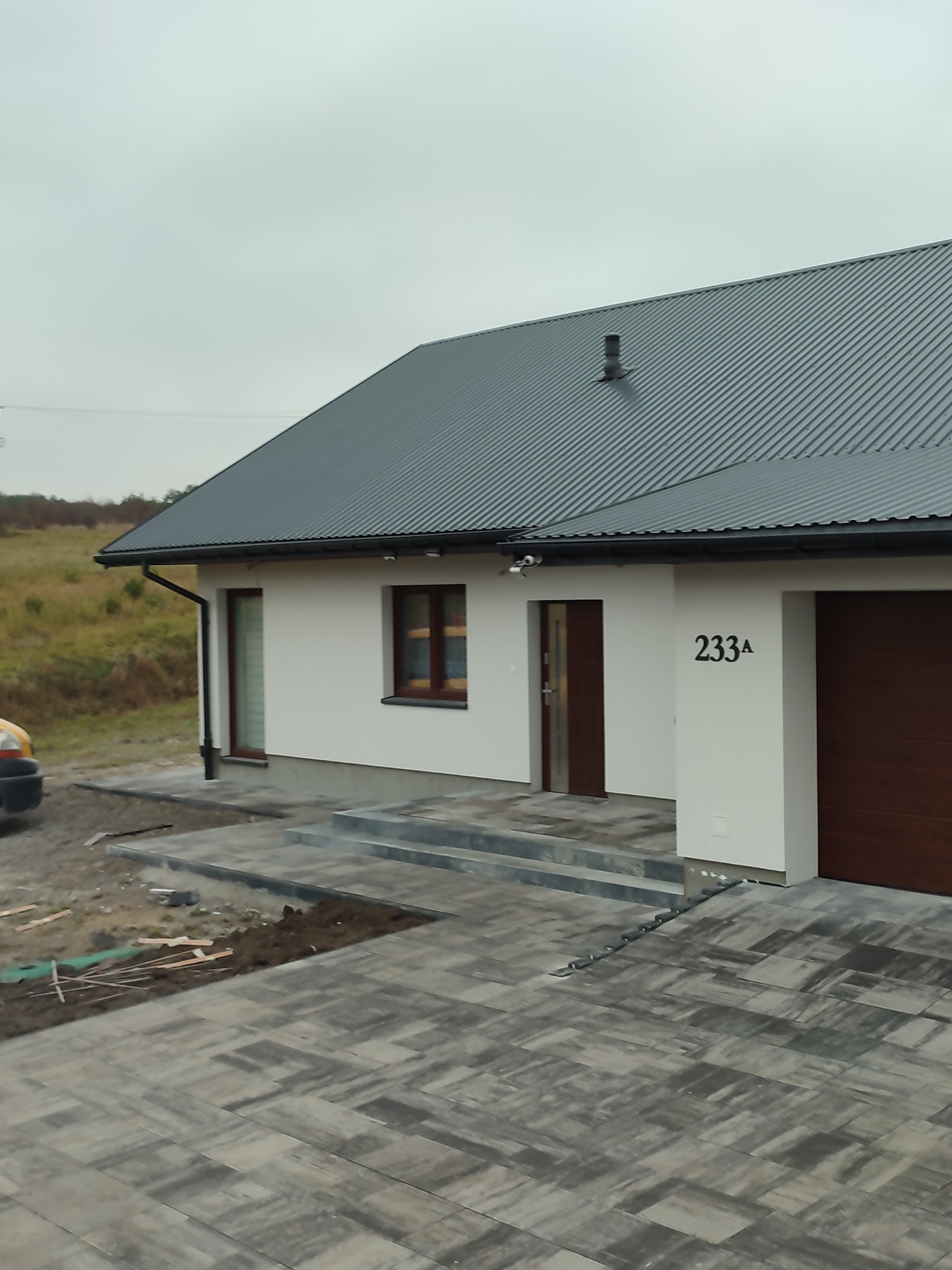 Nowoczesny dom jednorodzinny z szarą, metalową dachówką i świeżo ułożoną kostką brukową w odcieniach szarości, widoczne pozostałości po pracach ziemnych.