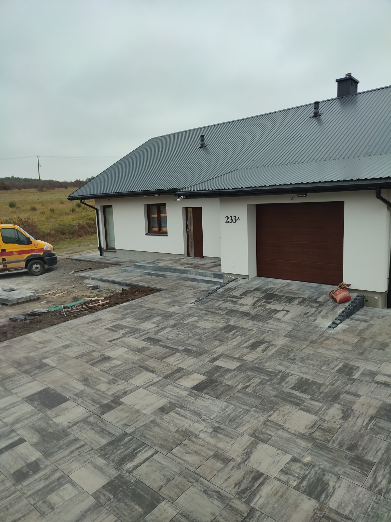 Nowoczesna posesja z jasną elewacją domu i nowo ułożoną, wzorzystą nawierzchnią z kostki brukowej w odcieniach szarości, z widocznym samochodem dostawczym w tle i workiem cementu przy garażu.